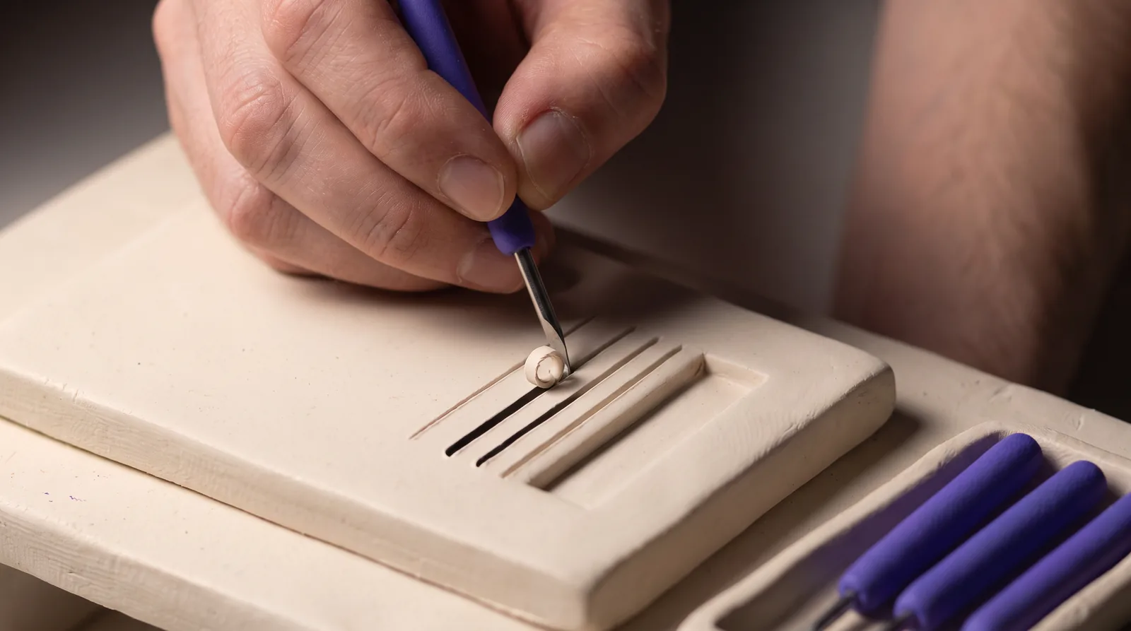 A human hand cuts deep lines into a thick clay slab.